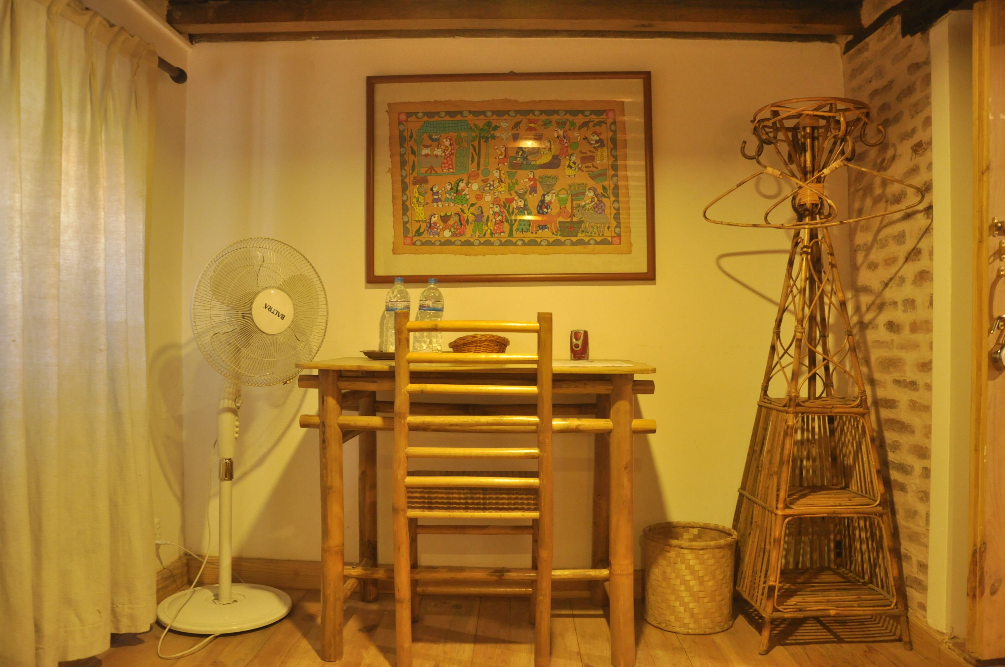 A room with a wooden desk, a standing fan, a wicker floor lamp, and a framed picture on the wall.