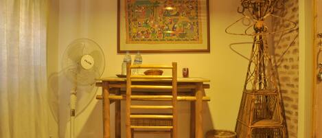 A room with a wooden desk, a standing fan, a wicker floor lamp, and a framed picture on the wall.
