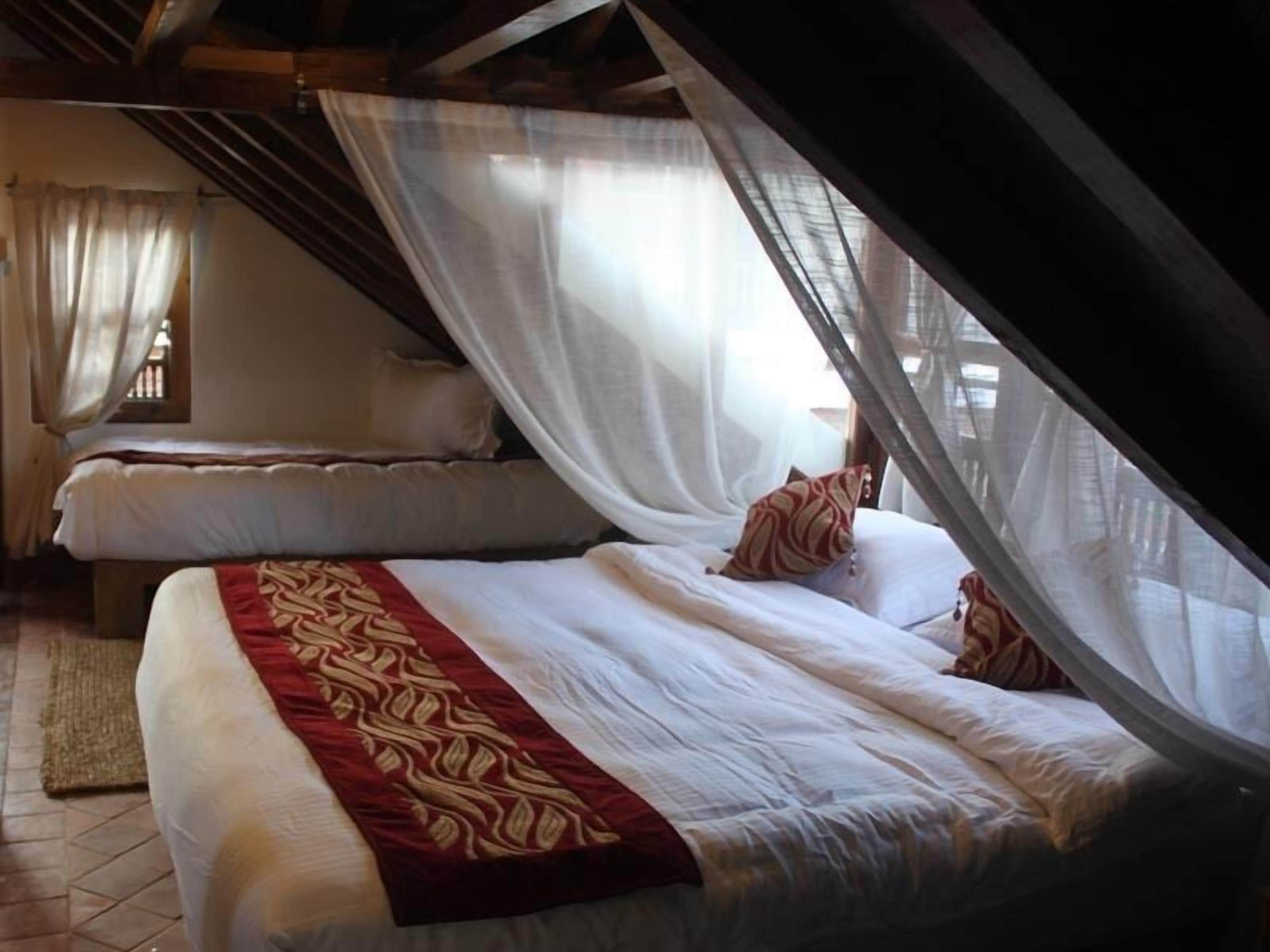 A bedroom with two beds, each covered with white linens and a red decorative border. The room has a sloped ceiling and a window with curtains.