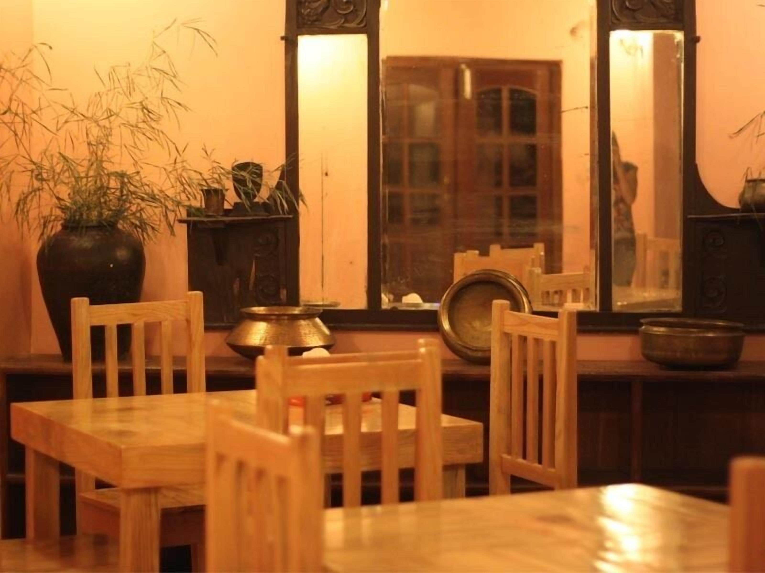 A wooden dining table with chairs, a large vase with plants, and a mirror reflecting a window with a view of a building.