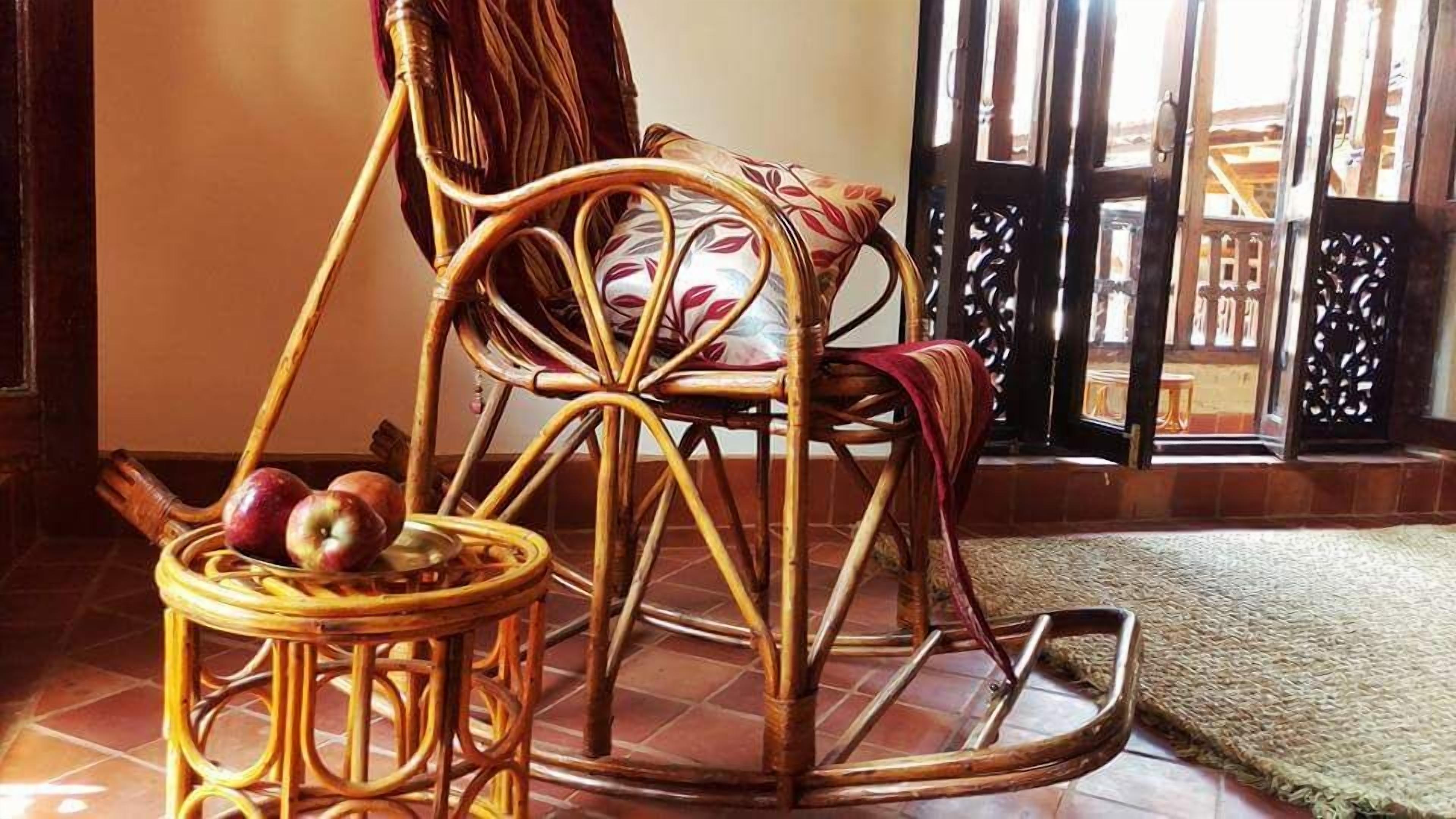 Un fauteuil à bascule en osier avec un coussin, une petite table ronde sur laquelle sont posées des pommes, et un tapis tissé sur un sol carrelé.