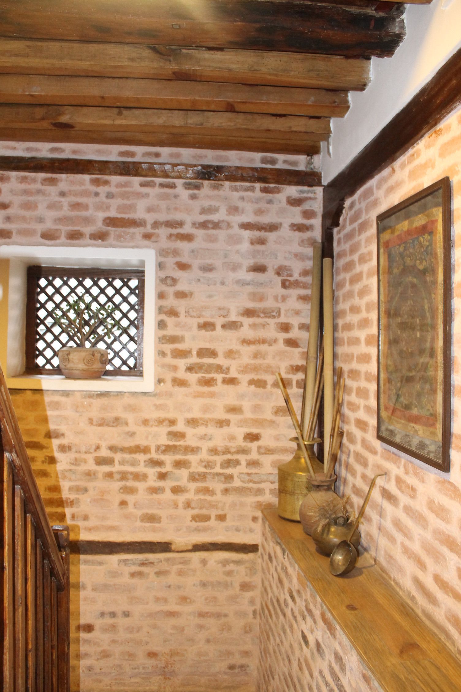 Une pièce rustique aux murs en briques apparentes, au plafond en bois, à la petite fenêtre ornée d’un treillis et à l’étagère où sont disposés des objets décoratifs.