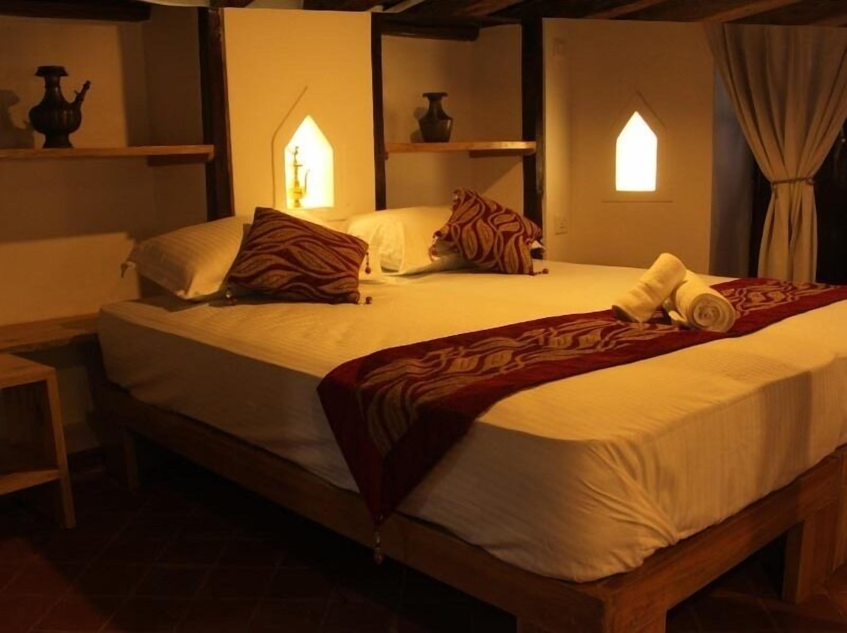 Una cama individual con estructura de madera, ropa de cama blanca y una manta de color rojo con estampado. Hay dos lámparas fijadas a la pared, con un diseño único, y una mesita de noche junto a la cama.