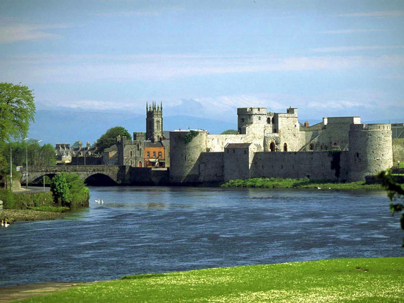 A historic stone castle with a river in the foreground and a bridge connecting to a town.