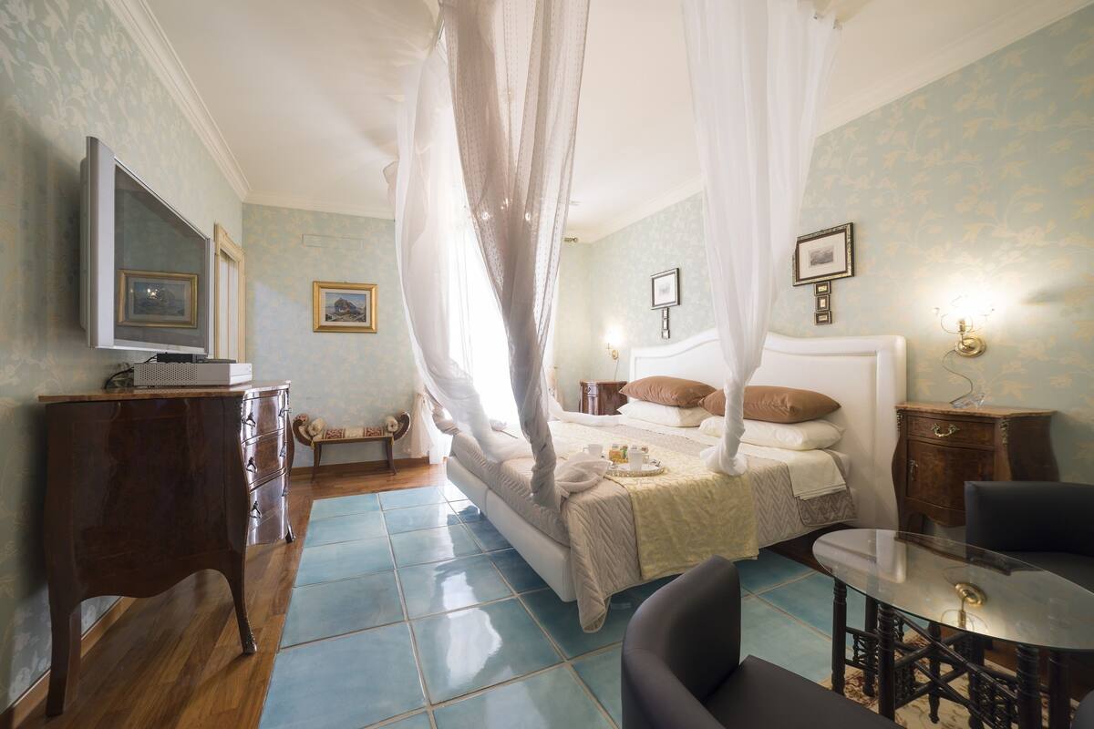 A bedroom with a four-poster bed, a bedside table, a chair, a television, and a small cabinet.