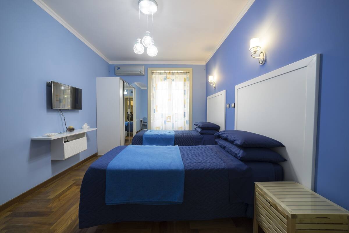 A bedroom with two beds, a TV, a bedside table, a wooden chest, and a window with curtains.