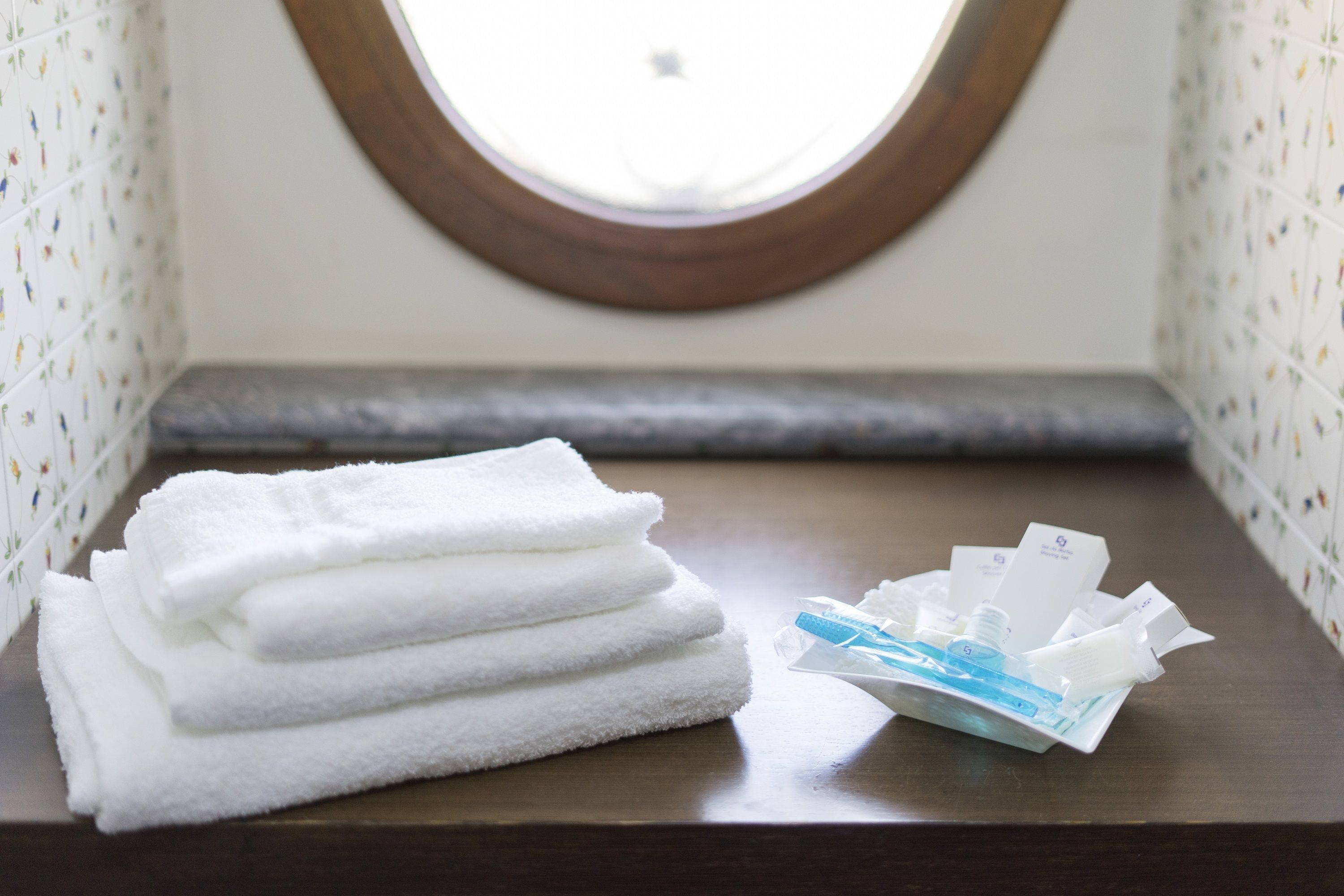 A stack of white towels and a package of toiletries on a wooden surface in a room with patterned wallpaper and a round window.
