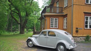 Une voiture ancienne est garée devant une maison en bois dotée d’un balcon.