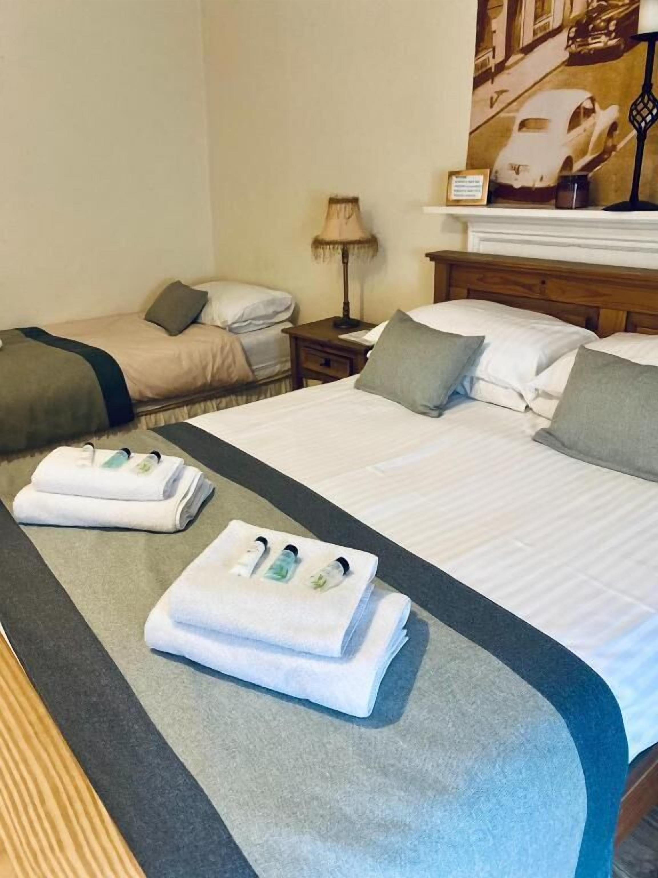 A hotel room with two beds, a nightstand, a lamp, and a wall-mounted clock.