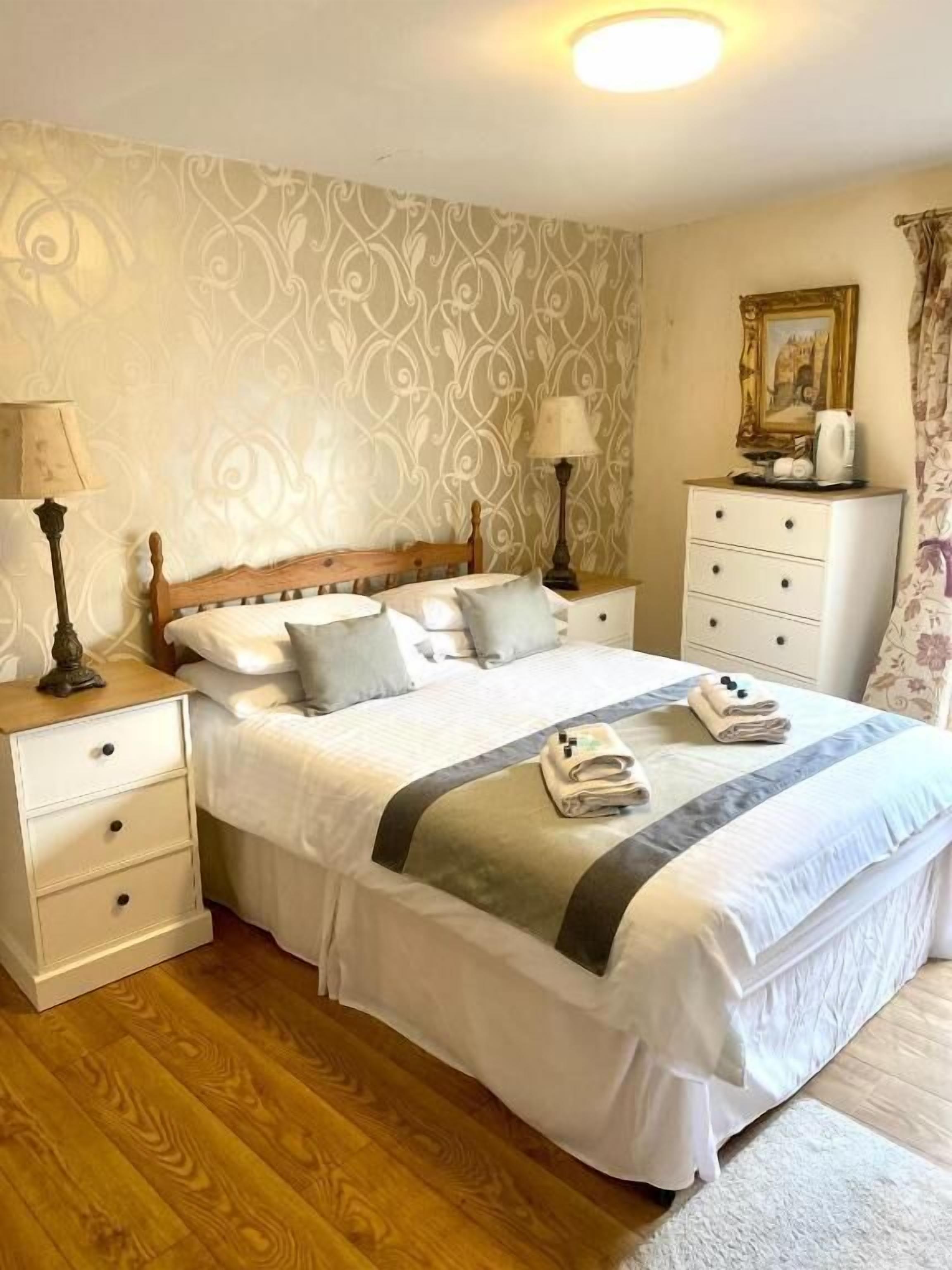 A bedroom with a wooden bed, white nightstands, a dresser, and patterned wallpaper.