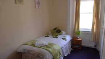 A small, neatly made bed with a green blanket and a white pillow, a bedside table with a green lamp, and a window with curtains.
