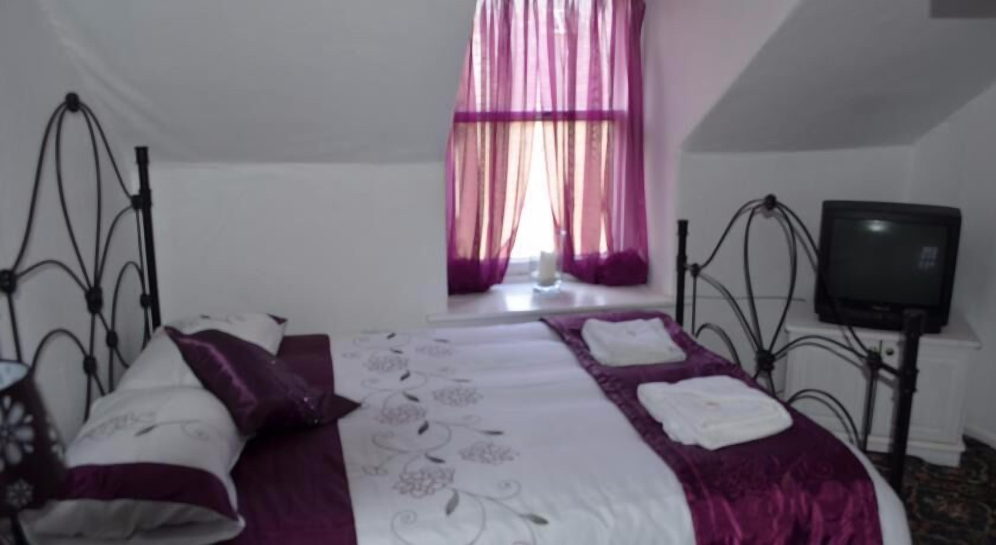 Un lit bien fait, recouvert d’une couvre-lit violette et blanche, un téléviseur posé sur un meuble à droite, et une fenêtre ornée de rideaux violets.