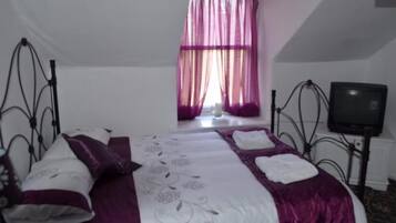 Un lit bien fait, recouvert d’une couvre-lit violette et blanche, un téléviseur posé sur un meuble à droite, et une fenêtre ornée de rideaux violets.