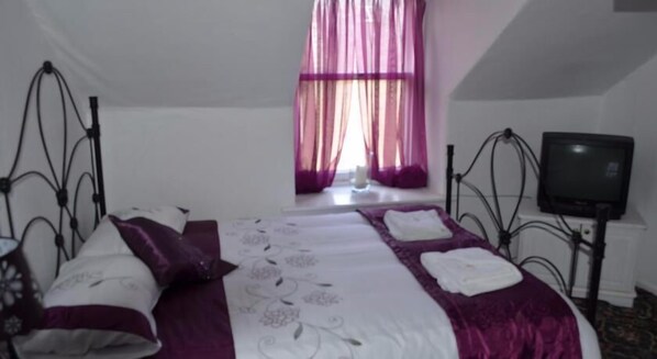 A neatly made bed with a purple and white bedspread, a television on a stand to the right, and a window with purple curtains. - Newcastle Arms Hotel (Coldstream)