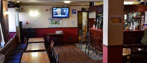 A restaurant interior with wooden tables and chairs, a television mounted on the wall, and a bar area with bottles.