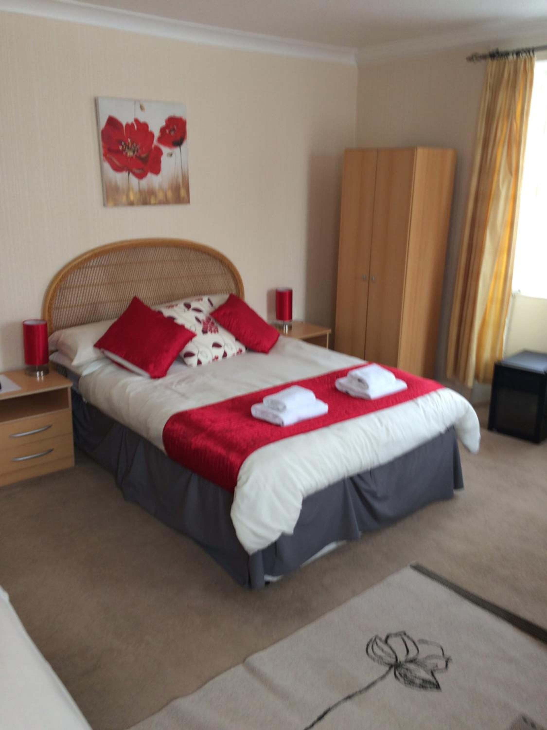 A neatly made bed with red and white bedding, a wooden wardrobe, and a red flower painting on the wall.