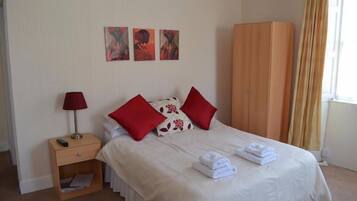 Una cama bien hecha con almohadones rojos, un armario de madera y una mesita de noche con una lámpara.