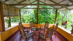 Un espacio al aire libre con techo, amueblado con sillas y mesa de mimbre, rodeado de abundante vegetación.