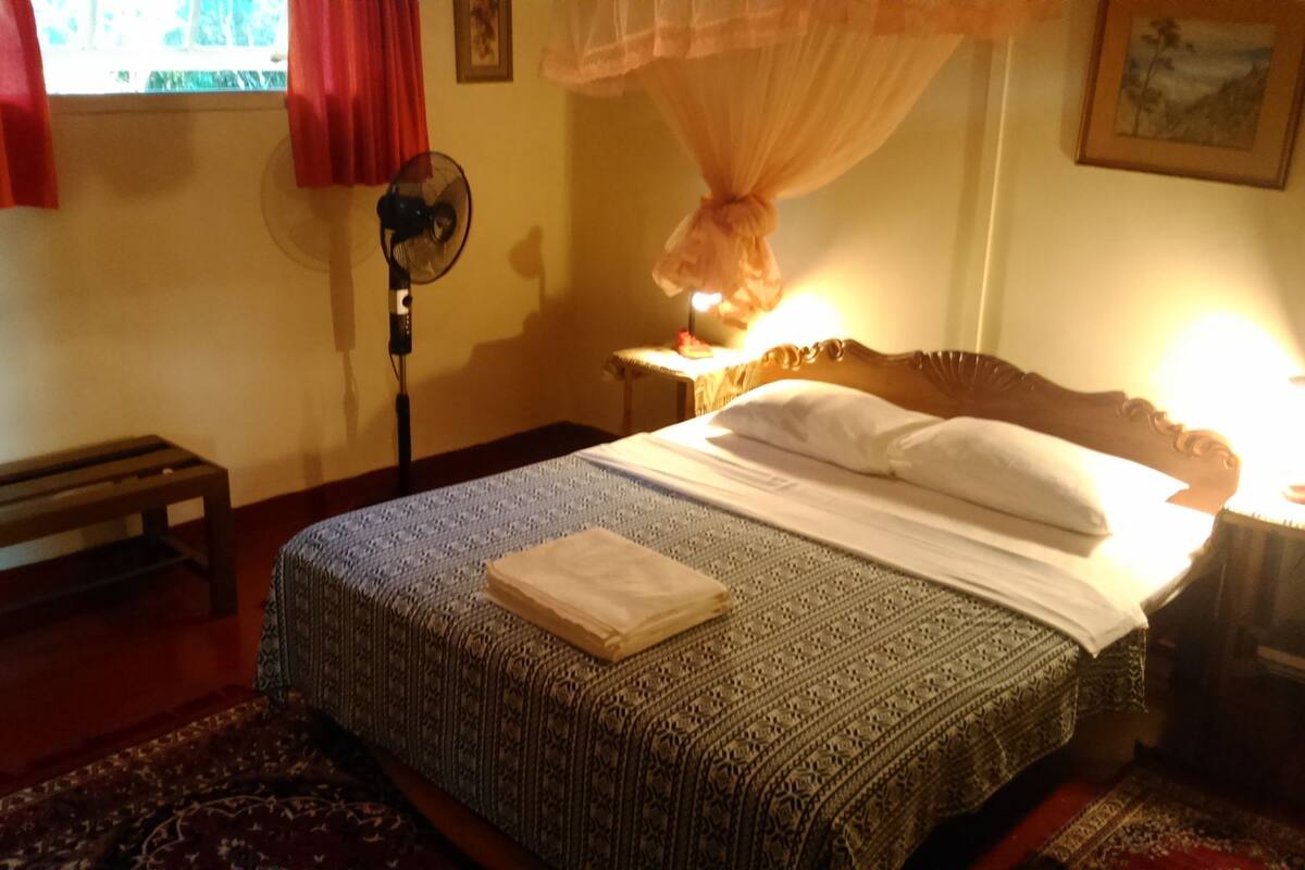 Ein ordentlich bezogenes Bett, ein gefaltetes Handtuch, ein Standventilator, eine Holzbank und ein Tisch mit einer Lampe.