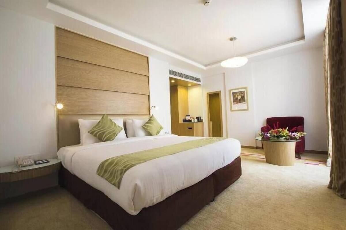 Quarto de hotel com uma cama grande, uma mesinha de cabeceira, uma cadeira vermelha, uma pequena mesa com flores e uma porta que dá para outro cômodo.