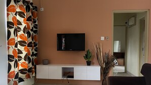 Un saló amb un televisor de pantalla plana fixat a un moble blanc, un sofà de color fosc, una taula de centre i una planta en test.