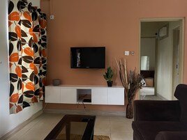Uma sala de estar com uma televisão de tela plana presa a um móvel branco, um sofá escuro, uma mesa de centro e um vaso com planta.