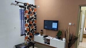 Un saló amb televisió penjada a la paret, una finestra amb cortina, una taula petita amb un bol i una planta en test.