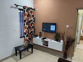 Uma sala de estar com televisão presa na parede, uma janela com cortina, uma mesinha com uma tigela e uma planta em vaso.