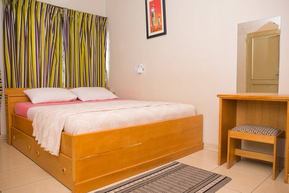A wooden bed with a pink and white bedspread, a wooden nightstand with a patterned cushion, a striped curtain, and a framed picture on the wall.