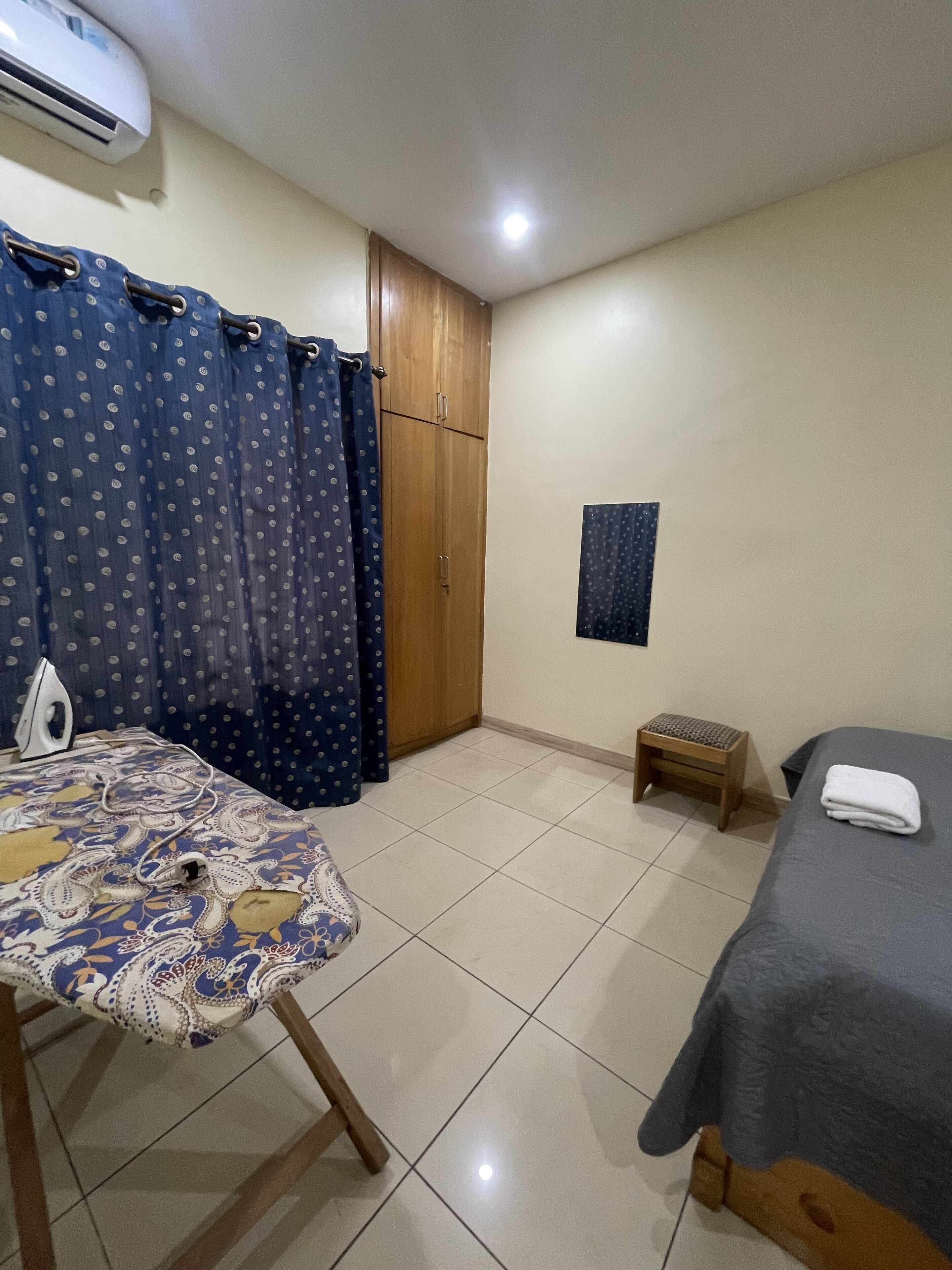 A bedroom with a bed, a small stool, a table with an iron, and a curtain.