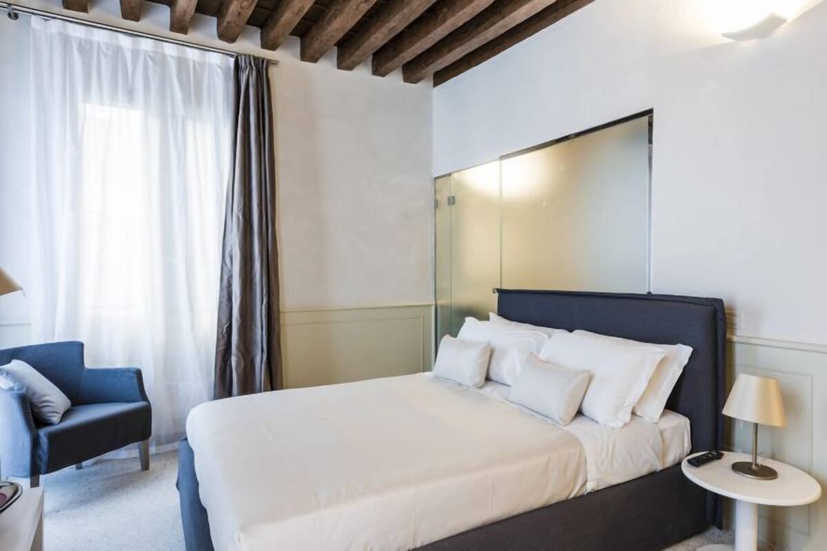Quarto de hotel moderno com uma cama grande, mesa de cabeceira com abajur, uma cadeira e um guarda-roupa.
