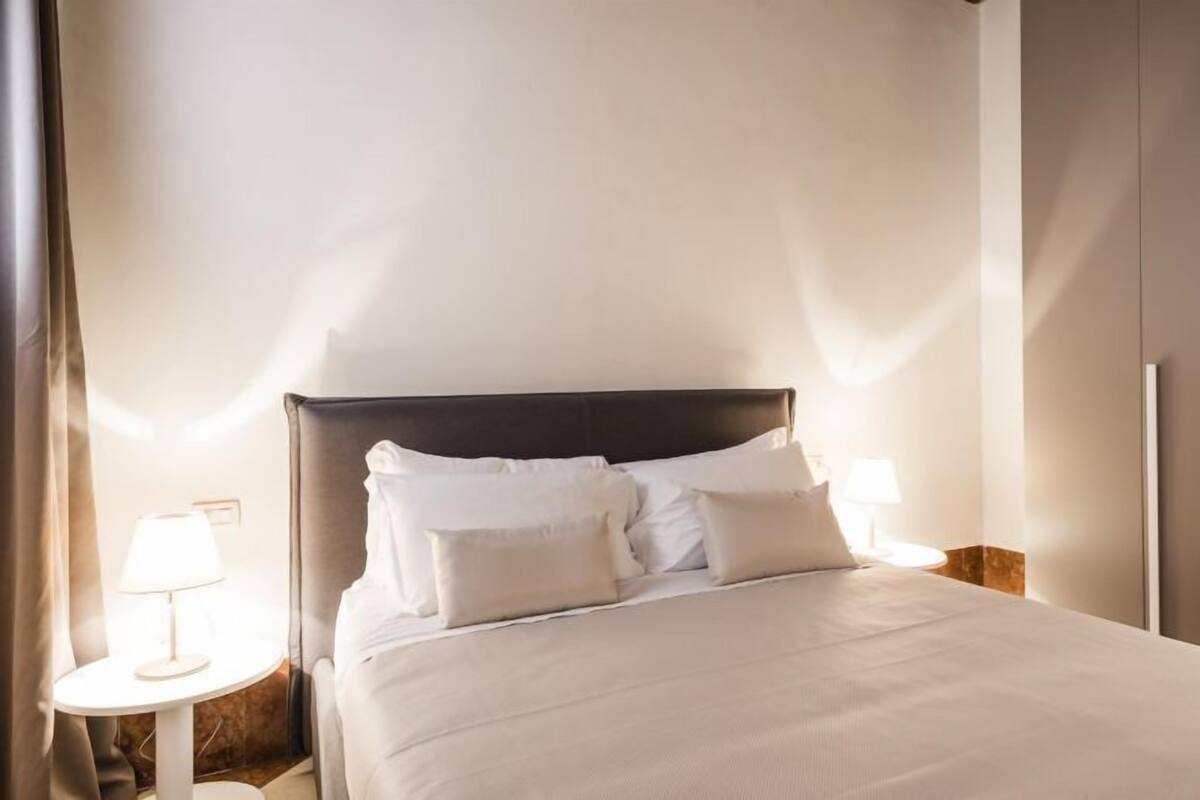 Uma cama bem arrumada com lençóis e almofadas brancos, uma luminária de cabeceira e um guarda-roupa em um quarto de hotel.