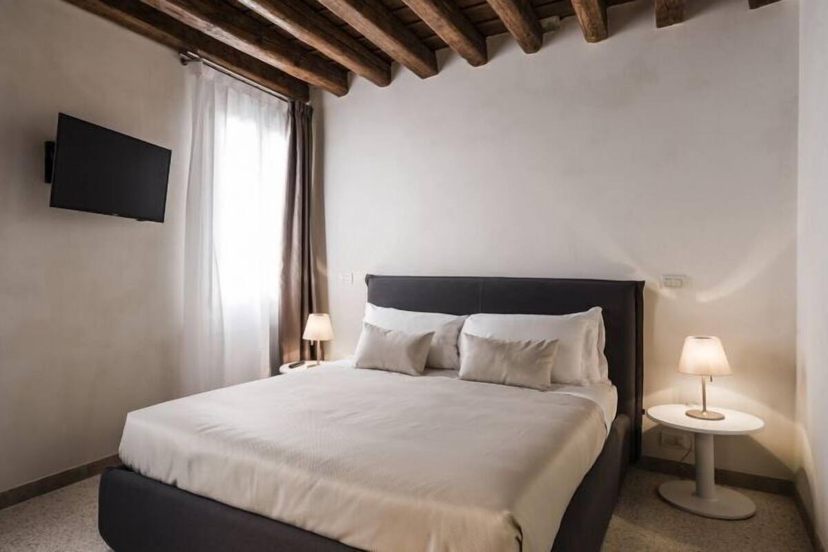 Quarto de hotel com uma cama, uma televisão na parede e duas luminárias de cabeceira.