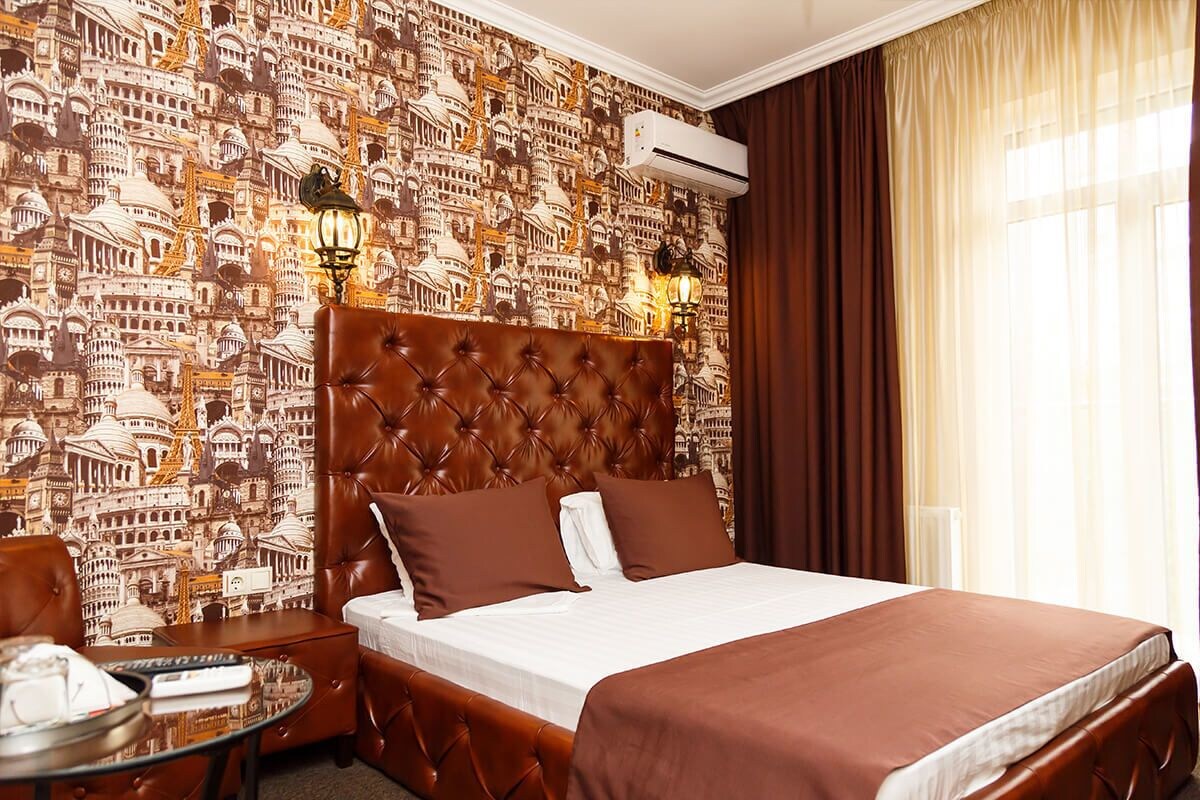 Une chambre d’hôtel avec un grand lit, une tête de lit, une table de chevet, une lampe et un mur recouvert d’un papier peint représentant un paysage urbain.