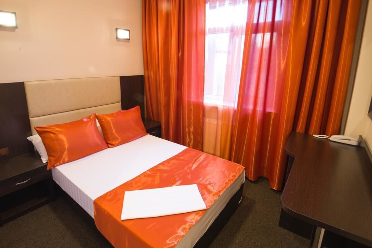 Une chambre d’hôtel avec un lit, une literie orange, une table de chevet avec un téléphone et des rideaux.