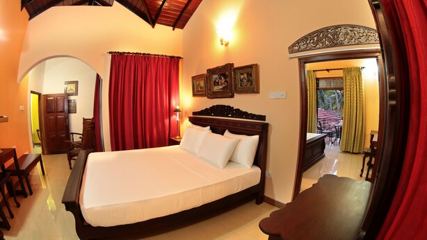 A hotel room with a large bed, a wooden headboard, a desk, and a chair. There is a door leading to a balcony with a view. - The Castle (Seeduwa - Katunayake)