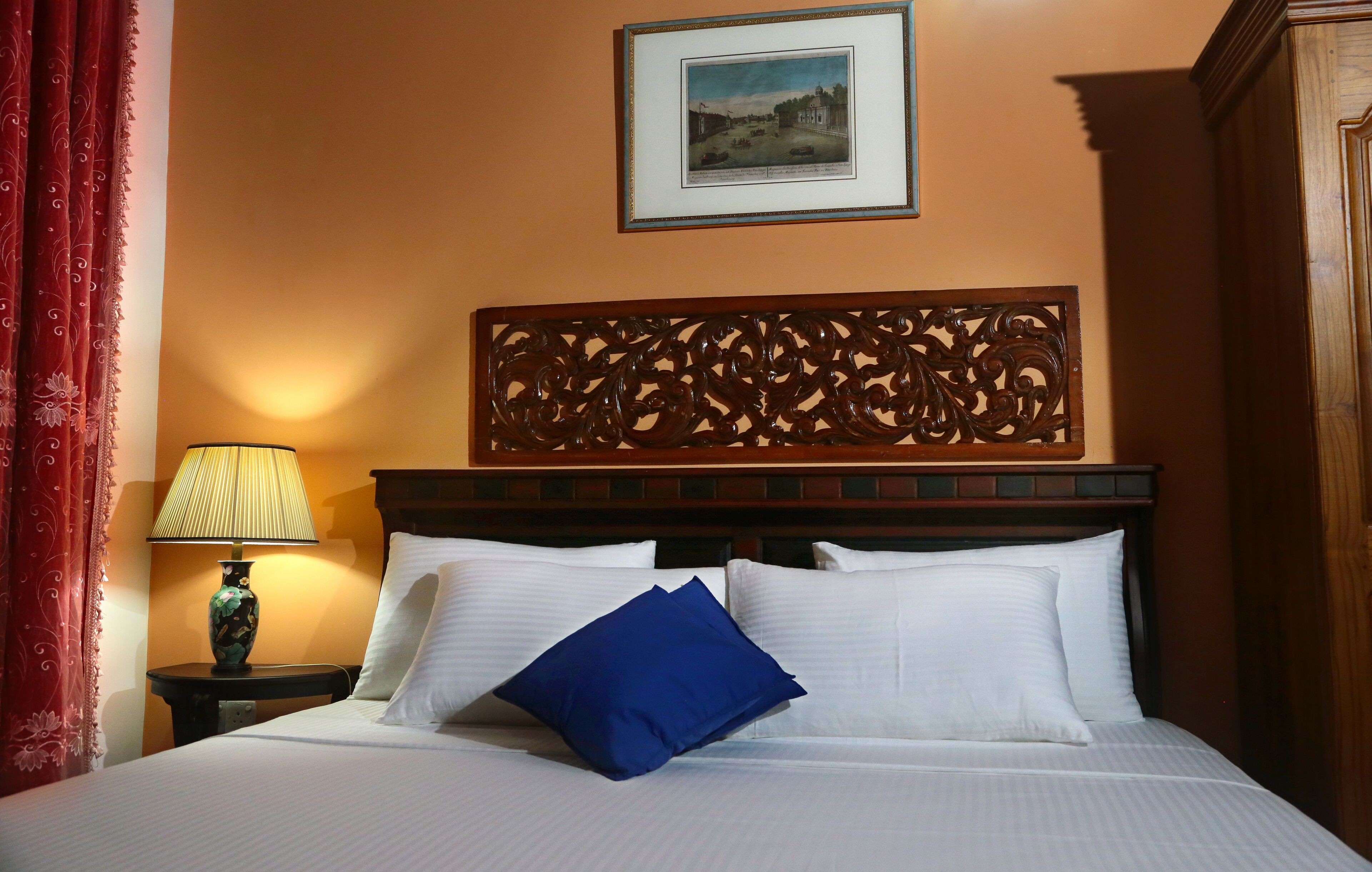 Una cama bien hecha con almohadas blancas y azules, un cabecero decorativo, un cuadro en la pared y una lámpara en una mesita de noche.