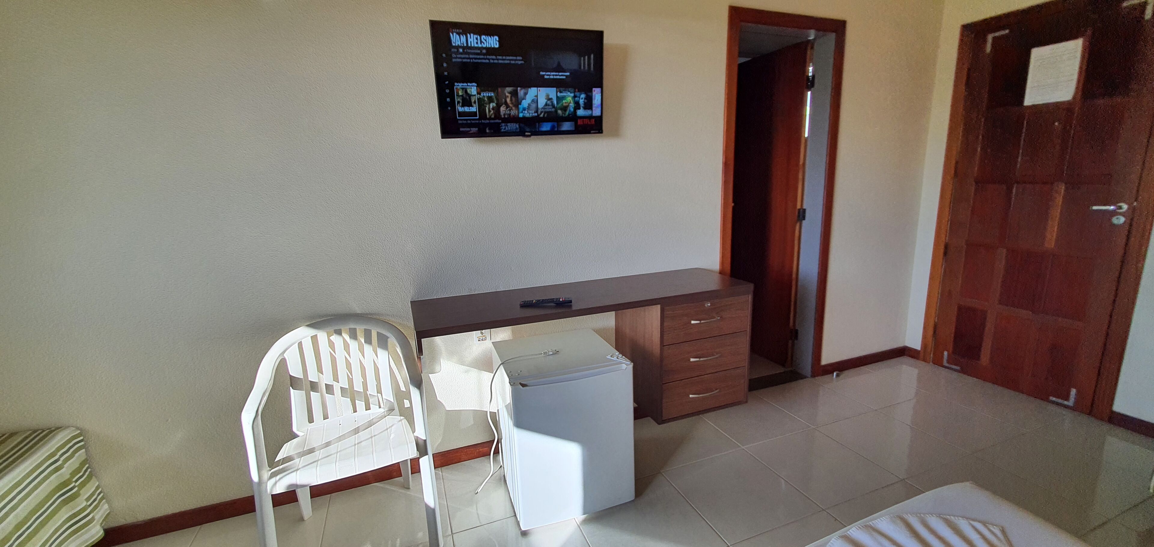 Um quarto pequeno com uma escrivaninha, uma cadeira, uma televisão na parede e uma porta.