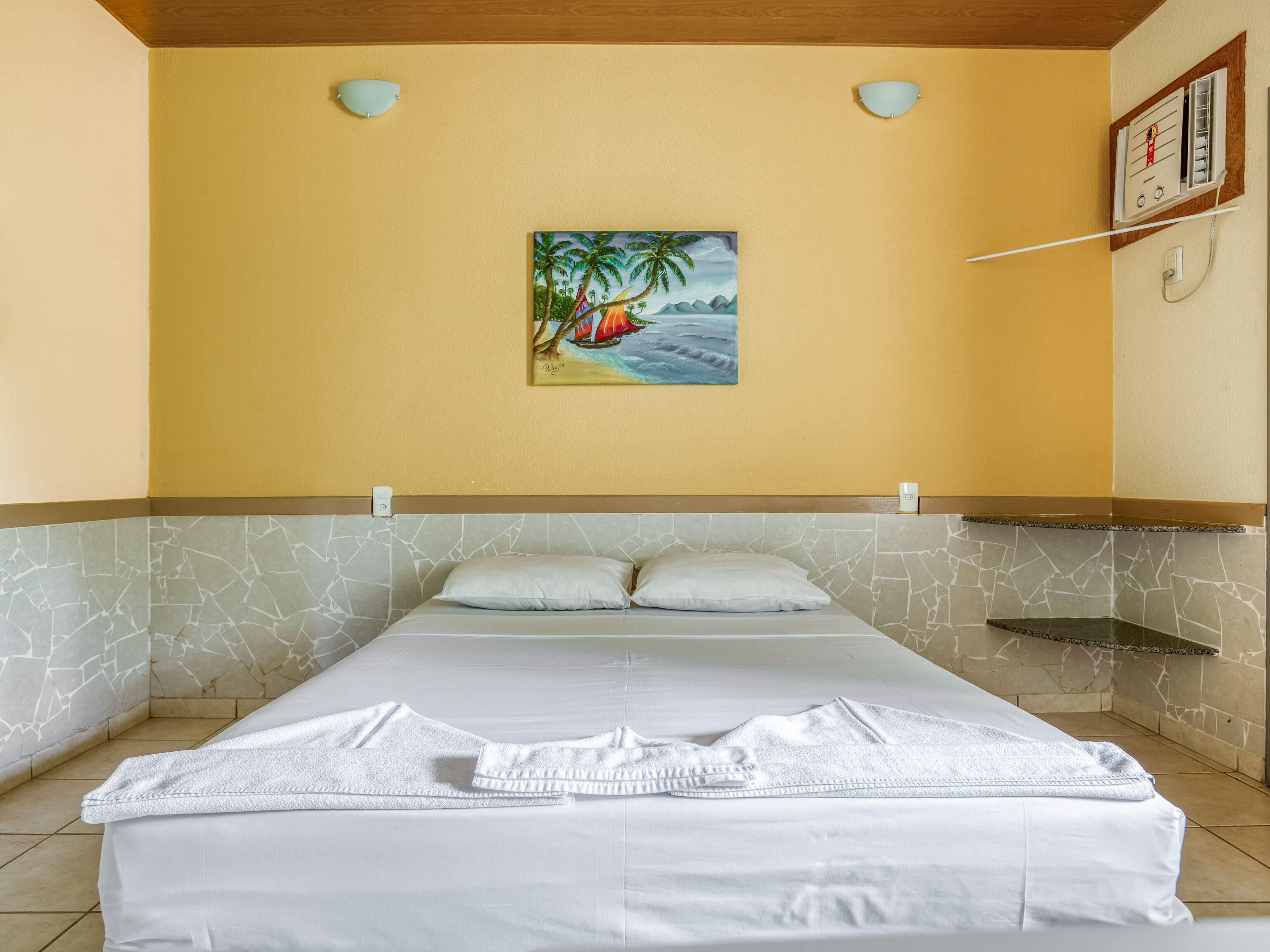 A neatly made bed with white linens and a towel, a painting of a beach scene on the wall, and a ceiling fan.