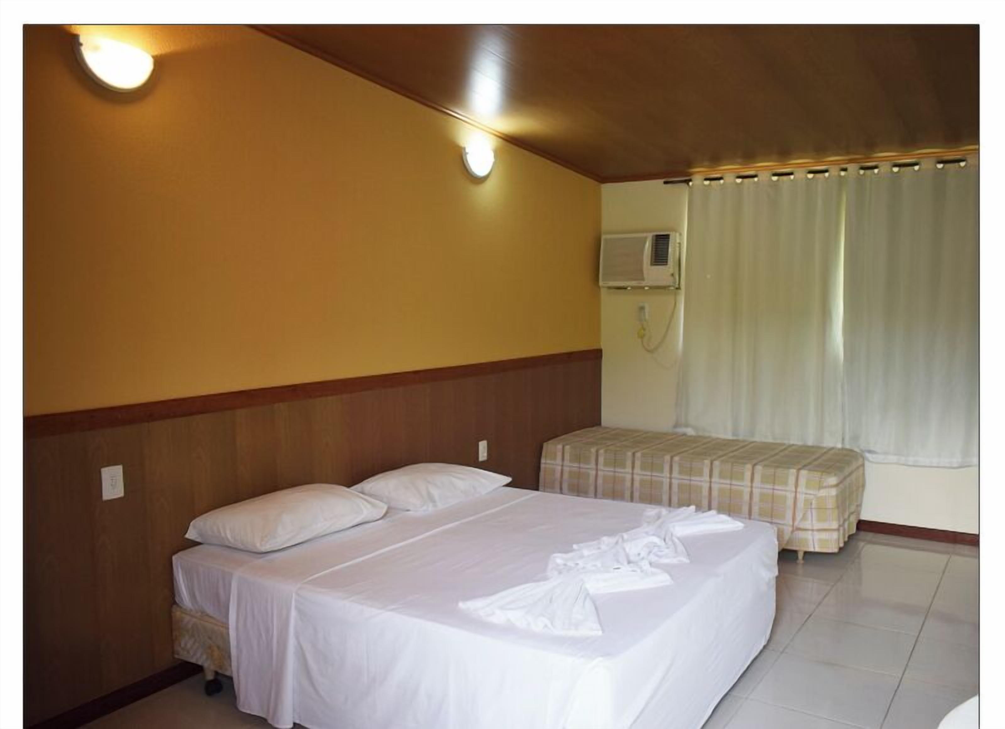 Habitación de hotel con cama, mesita de noche, una pequeña mesa, aire acondicionado de pared y ventana con cortinas.