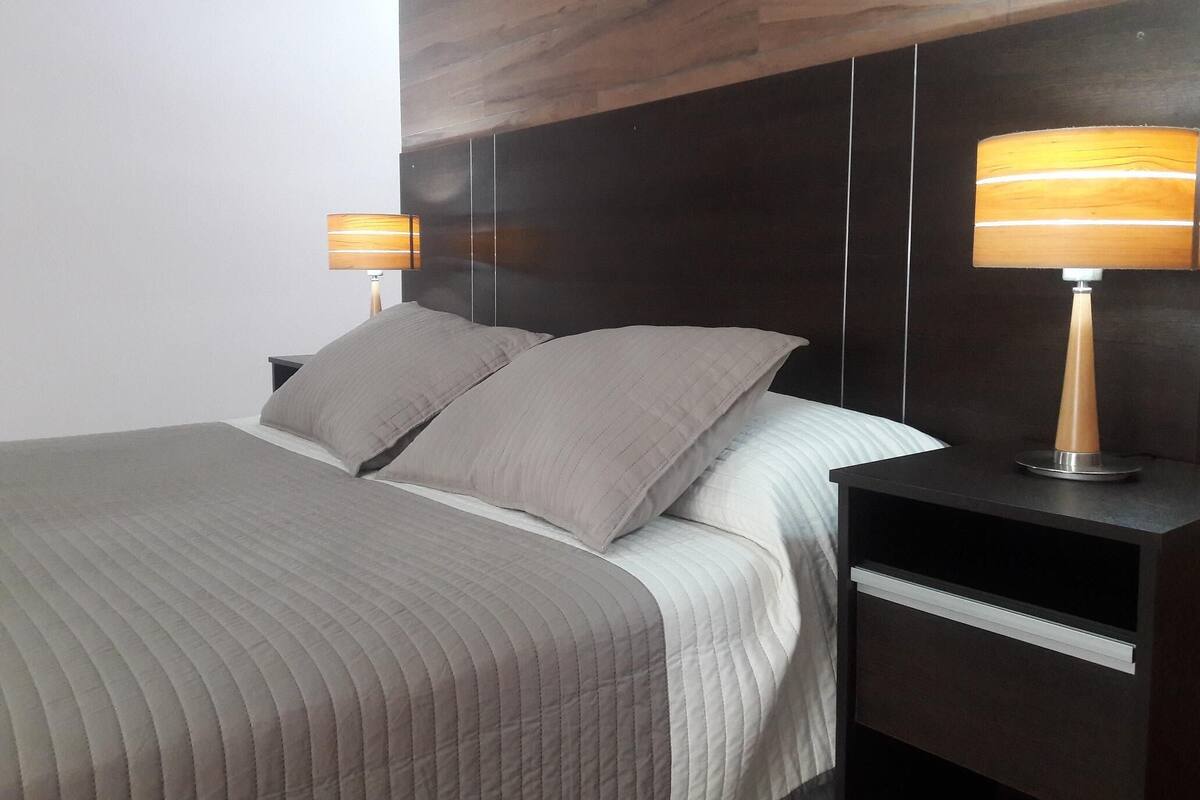 Una cama bien hecha con un edredón gris, dos lámparas de noche y un cabecero oscuro.