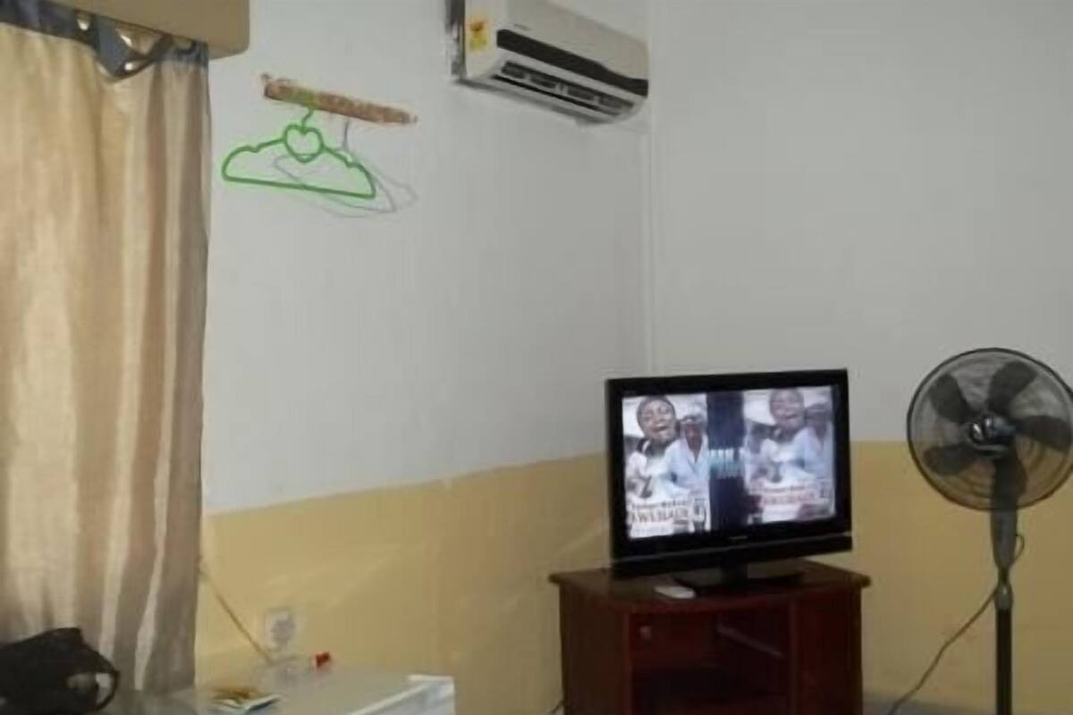 Une pièce avec un téléviseur posé sur un meuble en bois, un climatiseur blanc, un ventilateur et une petite table sur laquelle se trouve une télécommande.