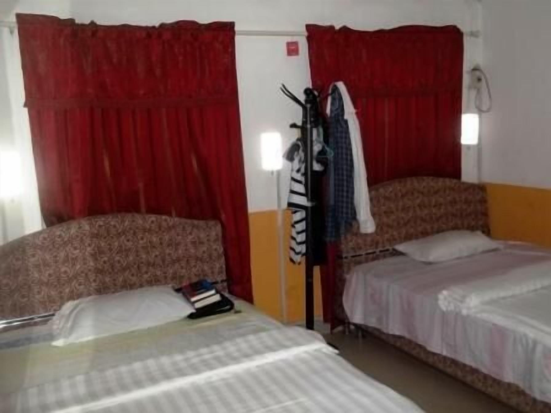 A room with two beds, one with a patterned headboard and the other with a plain headboard. There is a wardrobe with clothes hanging on it, and a small table with a book and a lamp.