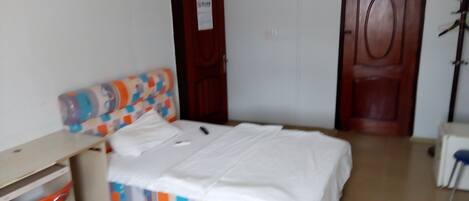 Una habitación pequeña y sencilla con cama, escritorio y silla. Hay dos puertas, una con un cartel, y un perchero en la esquina.