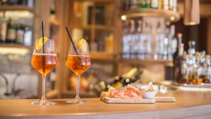 Two glasses of amber-colored drink with orange slices and straws on a wooden bar counter, with a bowl of sliced ham and crackers beside them. - Hotel Ambiez (Andalo)