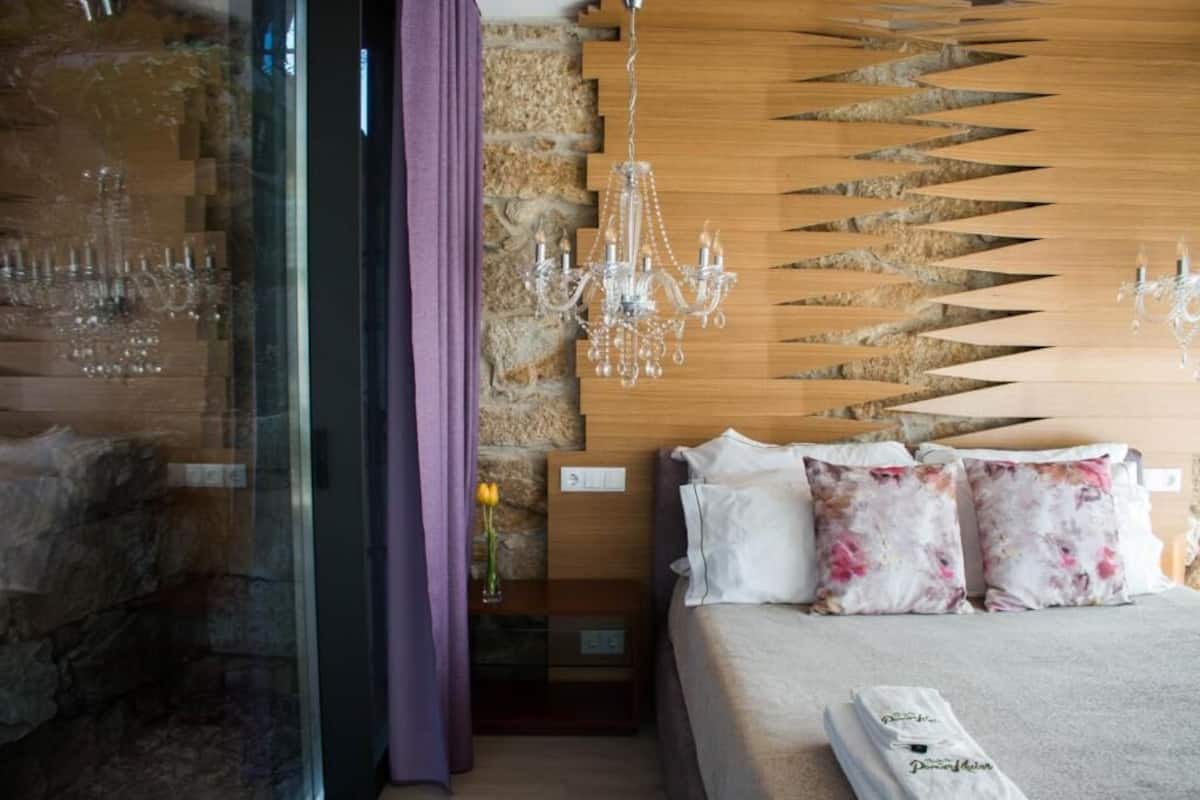Une chambre d’hôtel avec un lit, une tête de lit en bois, un lustre et une cloison en verre agrémentée de rideaux.