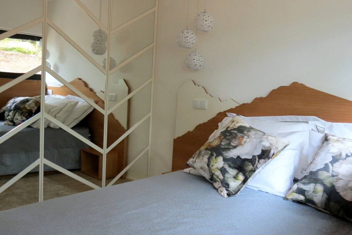 Une chambre à coucher avec un lit, des coussins à motifs floraux, une tête de lit en bois et un mur orné d’un motif géométrique.