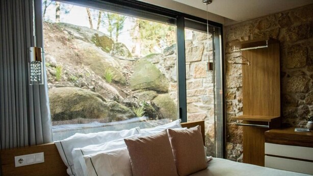 Un dormitorio con cama, almohadas, ventana con vistas a rocas y vegetación, un armario de madera y una pared de piedra.