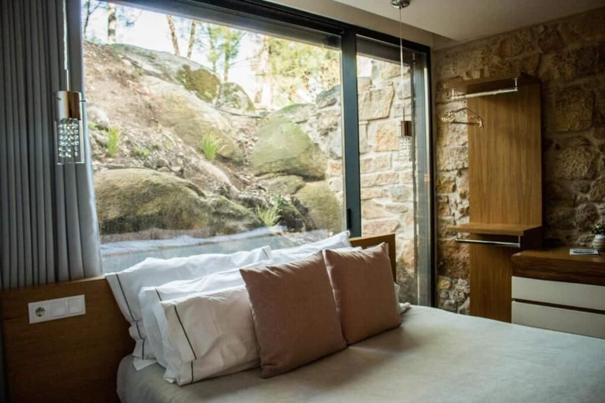Une chambre à coucher avec un lit, des oreillers, une fenêtre donnant sur des rochers et de la verdure, une armoire en bois et un mur en pierre.