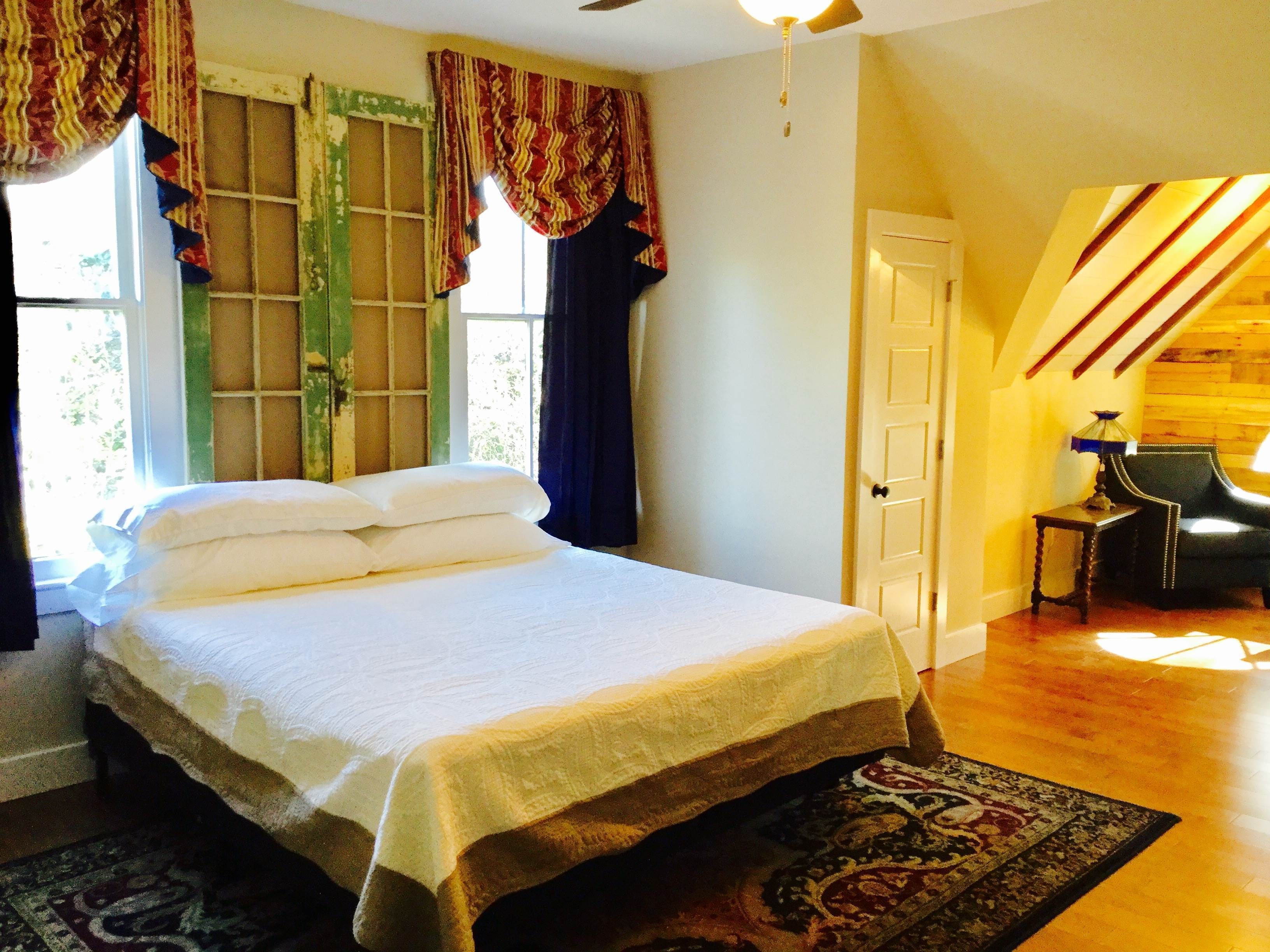 A bedroom with a bed, a window with curtains, a door, a chair, and a lamp.